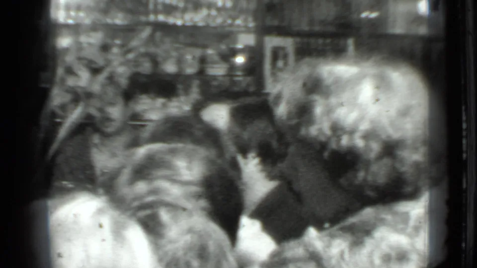 Grainy 8mm frame of adults at a crowded Moscow dinner table, with heads turned toward conversation and food partly visible.