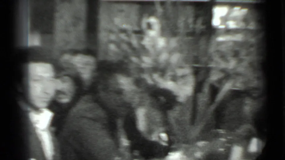 Soft black-and-white home-movie frame from the Moscow gathering, showing adults seated at a table with faces visible in a private room.