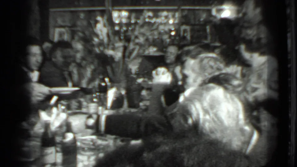 Grainy 1970s Moscow home-movie frame showing adults at a packed table, with serving plates, bottles, and decorative flowers in the room.