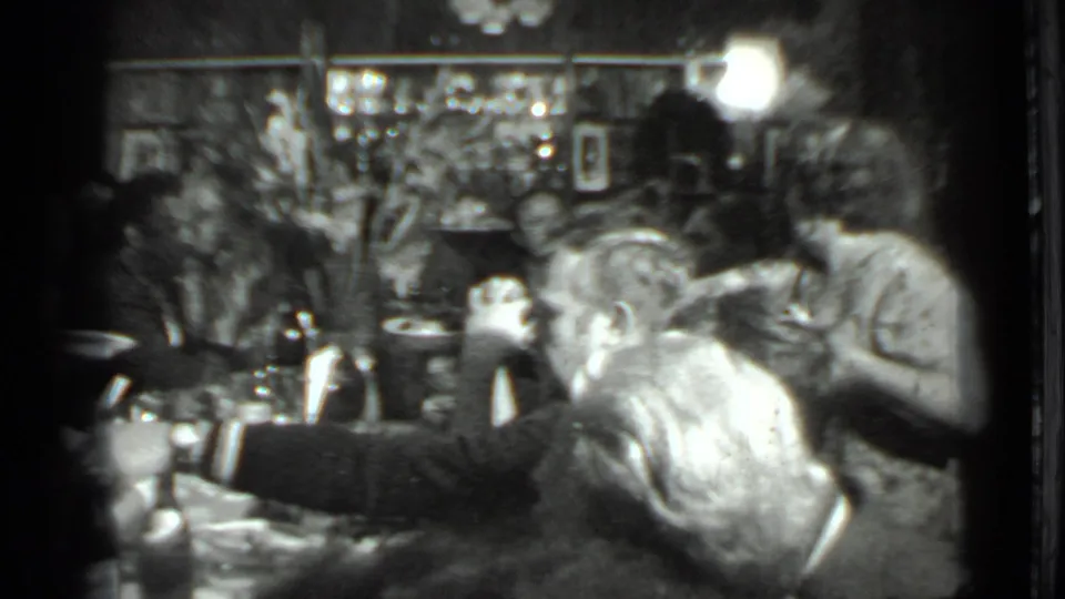 1975 Moscow home-movie frame showing adults gathered at a restaurant table with dishes, bottles, and wall decor in soft focus.