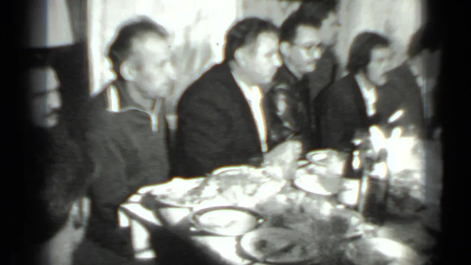 1975 Moscow home-movie frame showing adults in formal clothing seated at a long restaurant table with food and drinks.