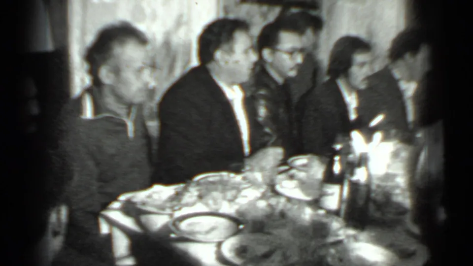 1975 Moscow home-movie frame showing adults seated closely around a dining table with plates, bottles, and glassware.