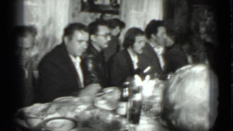Adults at a Moscow dinner gathering sit shoulder to shoulder around a table, with bottles, plates, and glassware visible.