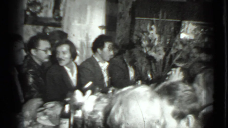 1970s Moscow home-movie frame of adults seated near a dining table, with bottles, plates, wall decor, and indoor furnishings visible.