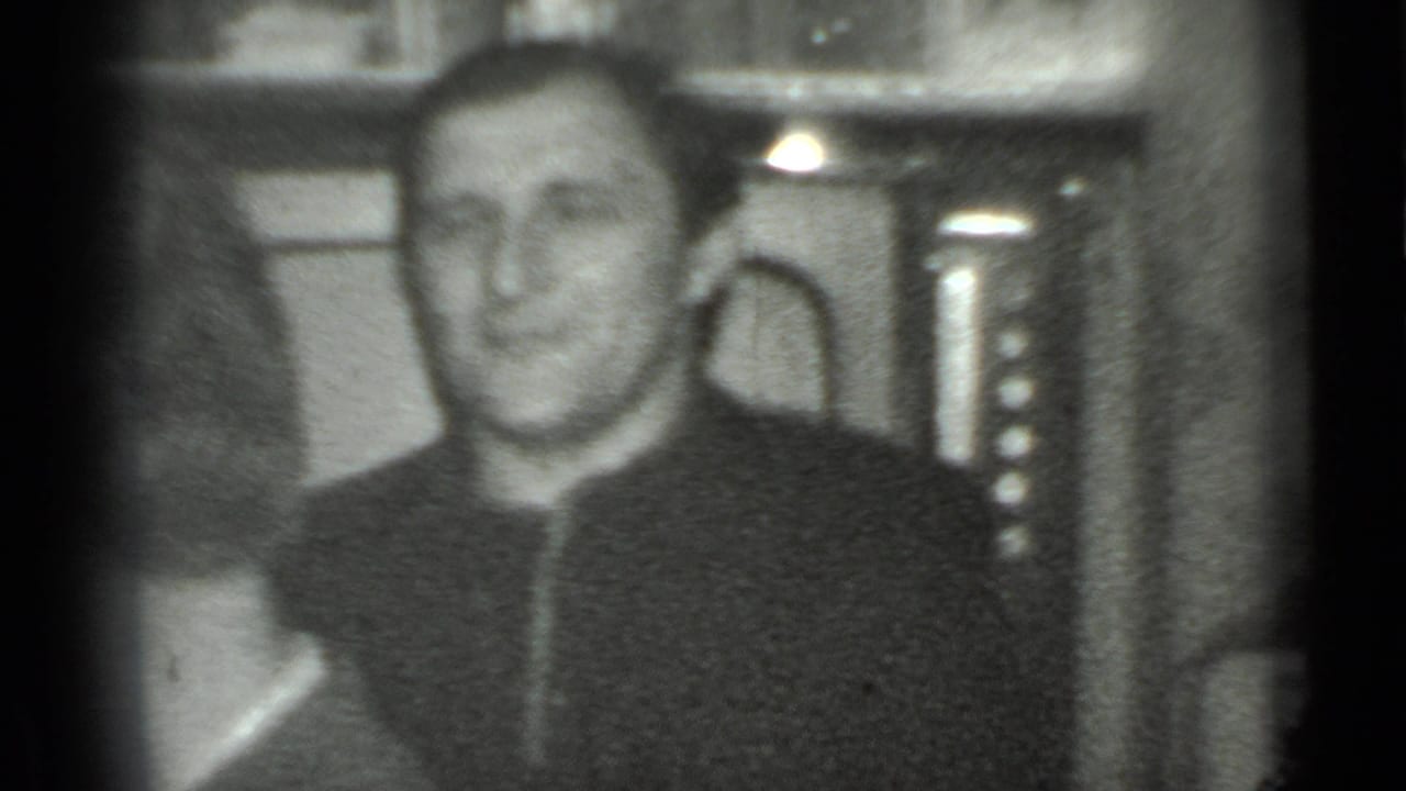 Man in a dark zippered jacket smiles toward the camera in a grainy indoor scene.