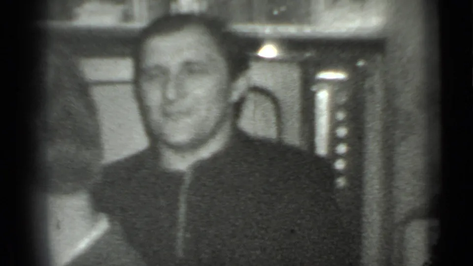 In 1975 Moscow home-movie footage, an adult man in a dark zippered jacket smiles toward the camera inside a private-looking room.