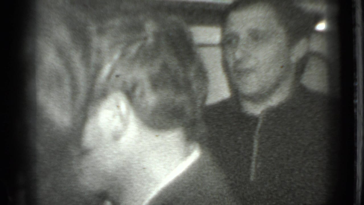 Two adult men face each other indoors, one seen from behind and one smiling at right.