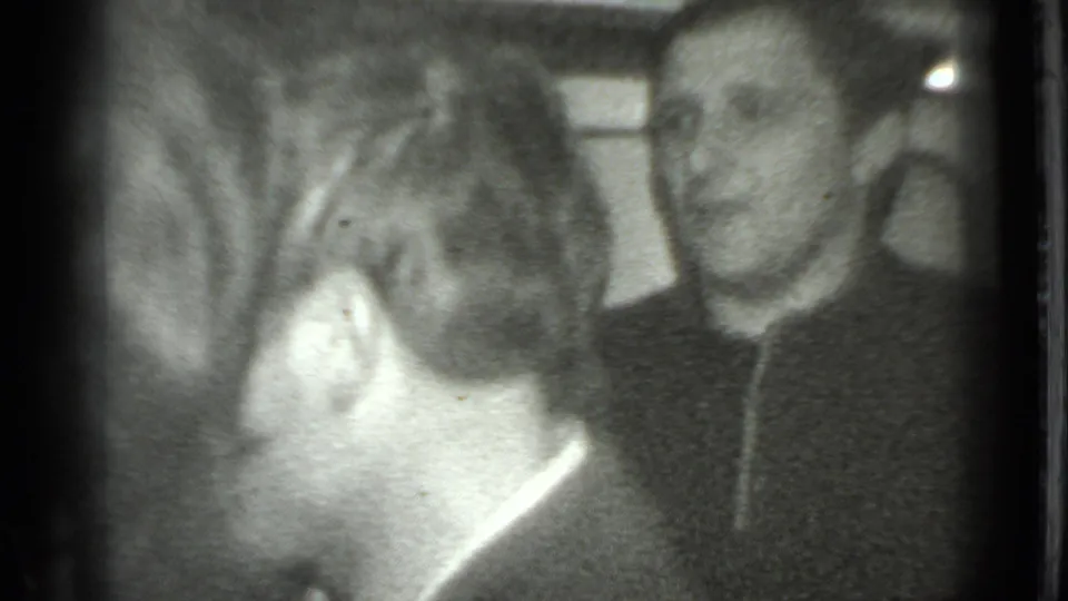 Two adult men converse indoors in the Moscow home-movie footage, with one man turned away and another smiling near the right edge.