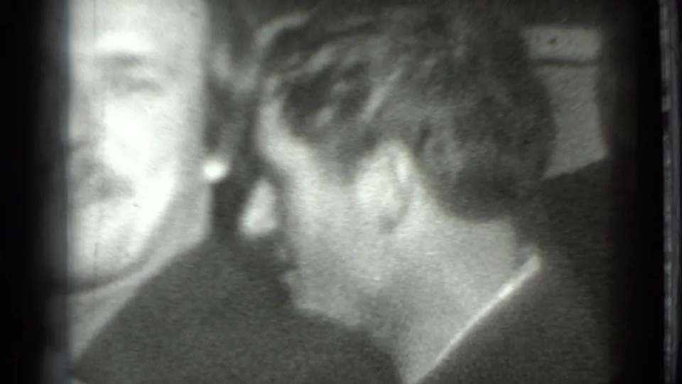 A close handheld frame shows two adult men in conversation, one partly cropped at left and the other seen from the back and side.