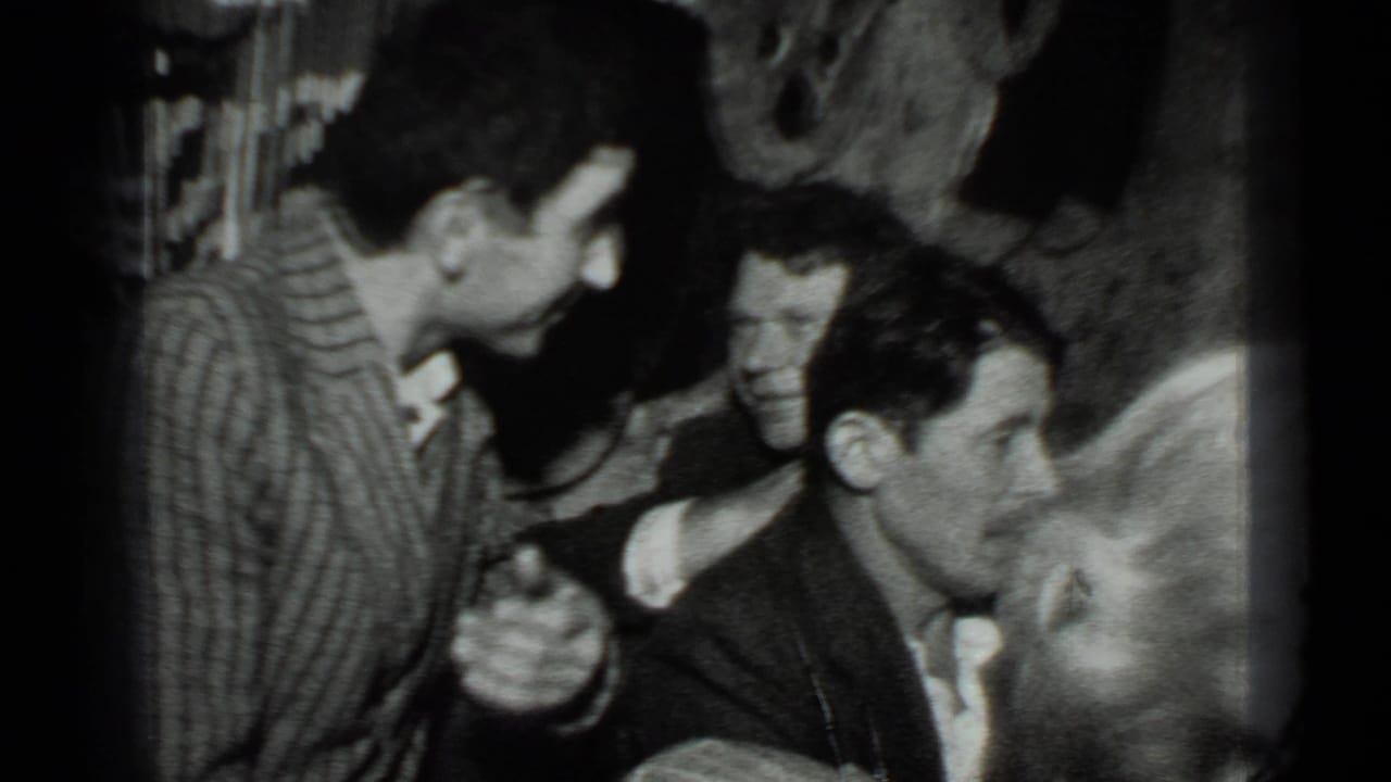 Several adults sit closely in a dim interior while one man in a striped shirt leans toward the group.