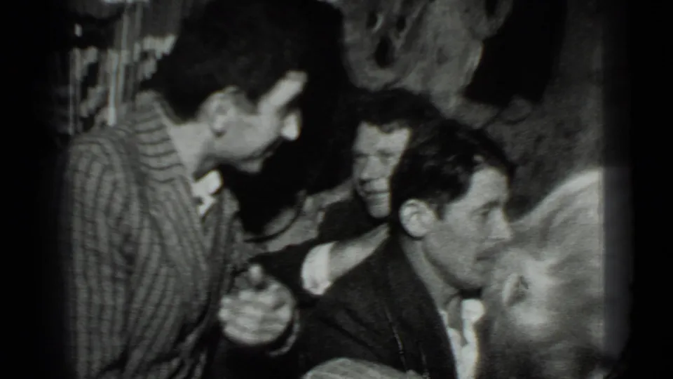 In a 1975 Moscow home-movie frame, adults gather closely indoors as a man in a striped shirt leans toward seated companions.