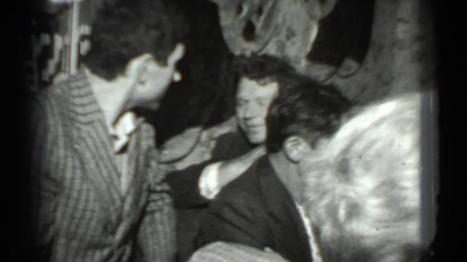 Adults appear in close conversation during a 1975 Moscow indoor gathering, with a striped shirt and patterned wall decor visible.