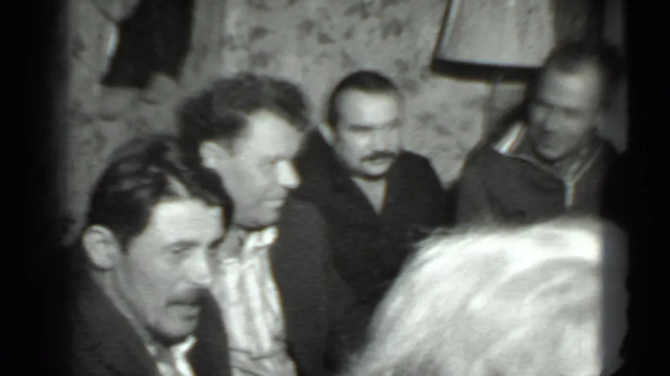 In 1975 home-movie footage, adult men appear to converse indoors in Moscow, framed tightly with soft grain and visible faces.