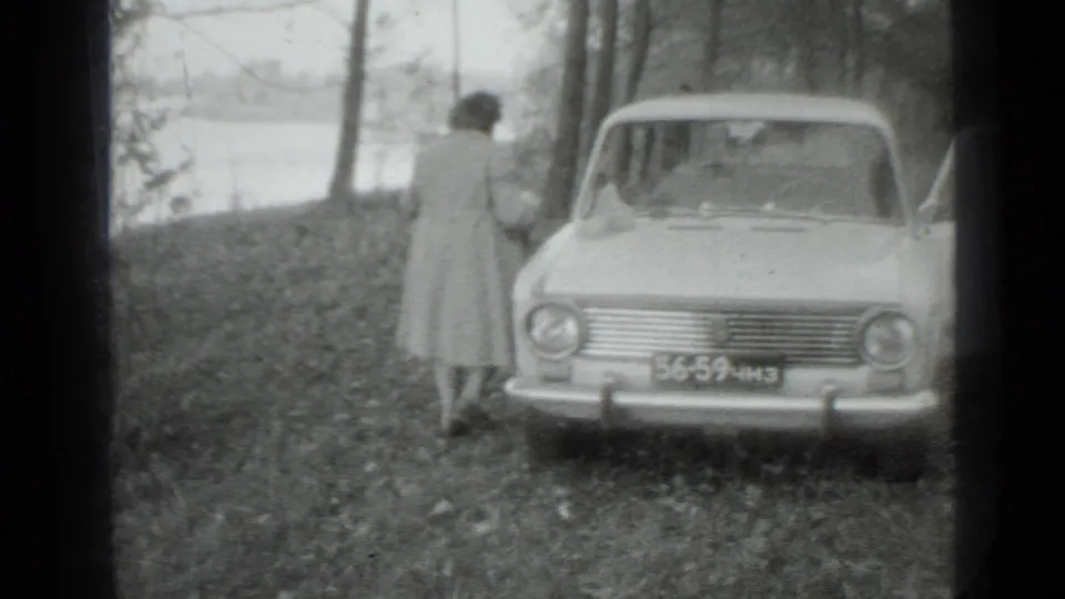 The woman faces a light-colored parked car near trees and a shoreline, captured in soft 8mm black-and-white footage.