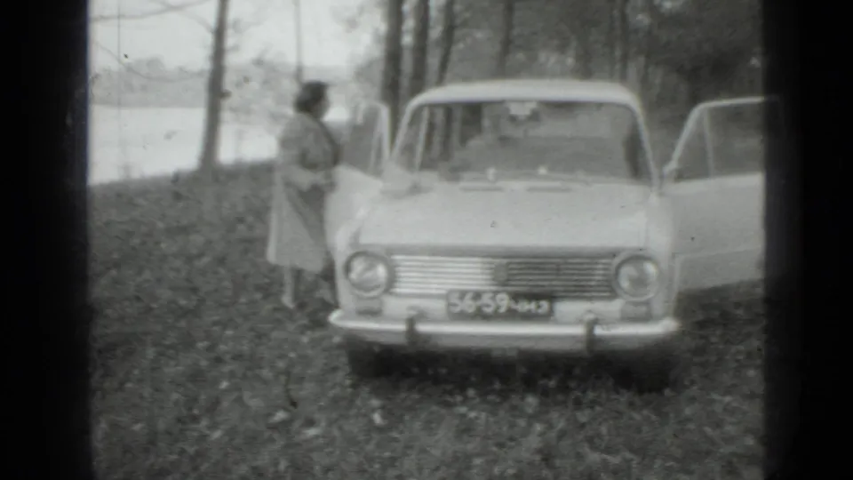 1975 Moscow-area home-movie frame showing a woman beside a light-colored sedan on a wooded slope near a lake.