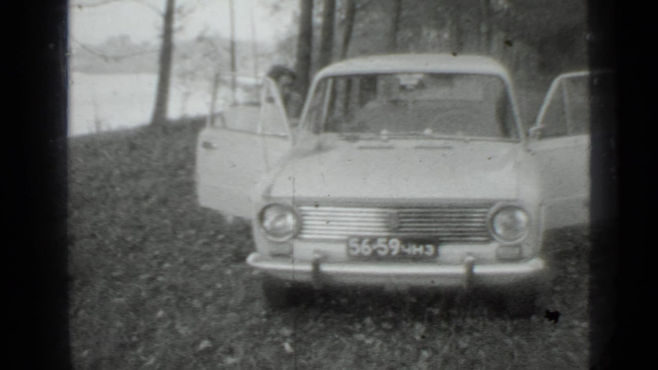 Front view of a parked sedan with both side doors open near a wooded lakeshore.