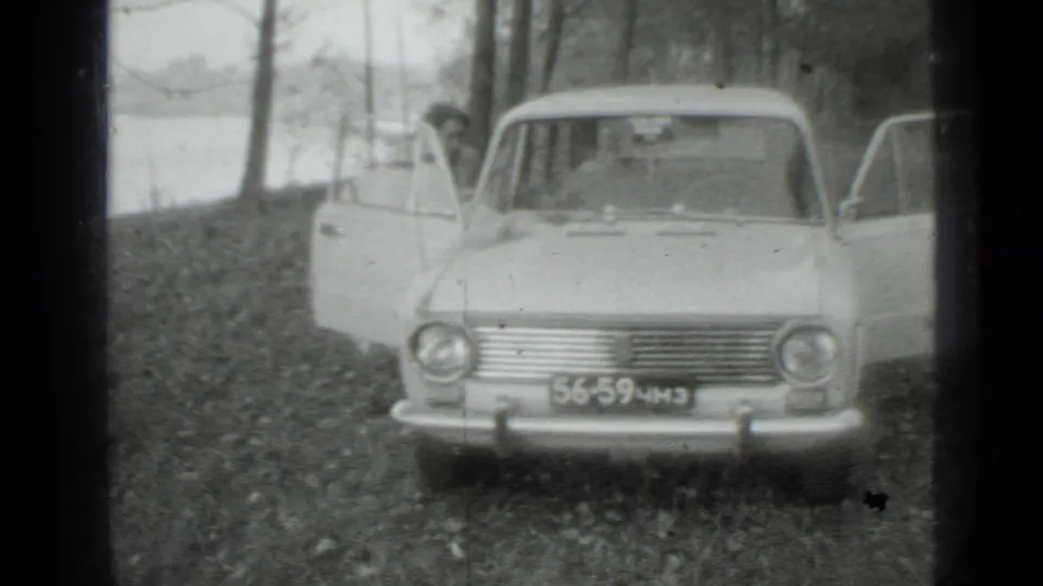 1975 Moscow-area frame of a light-colored sedan facing the camera with open doors on a wooded slope near water.