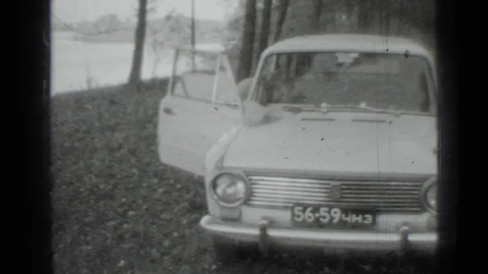 Closer 1975 Moscow-area home-movie view of the sedan front, open door, wooded bank, and lake beyond.