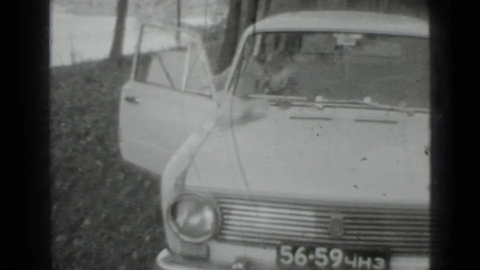 Close 1975 Moscow-area frame showing the front of a light-colored sedan and open door near a wooded lakeside.