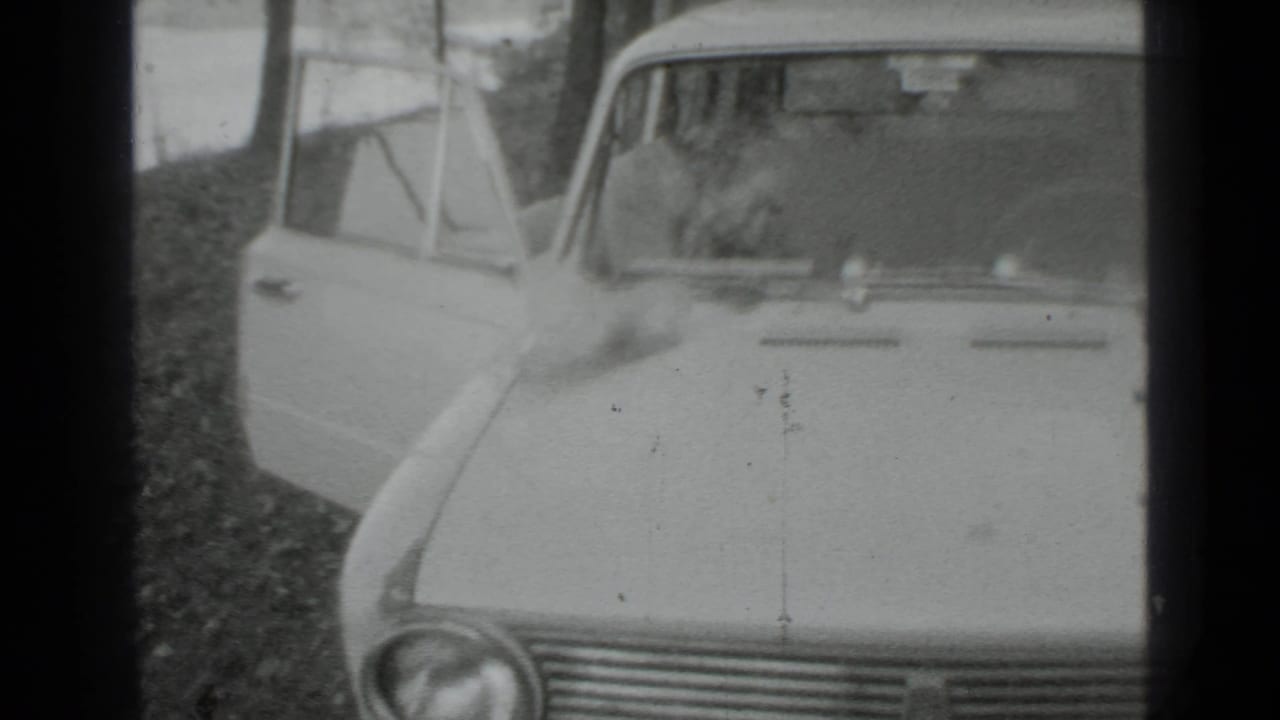 Front of a parked vintage car with its driver's door open beside trees and grass.