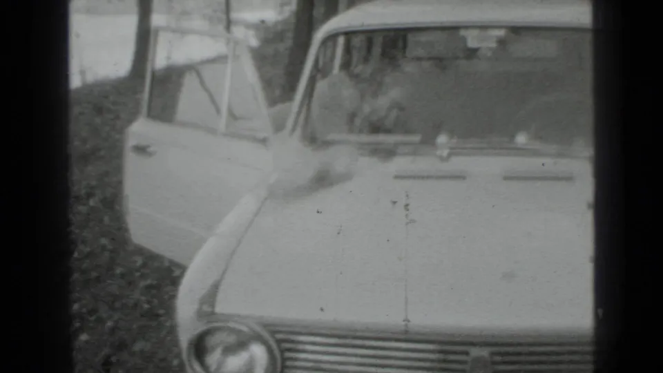 Black-and-white 8mm frame of a vintage car in Moscow, 1975, with the driver's door open in a quiet outdoor setting.