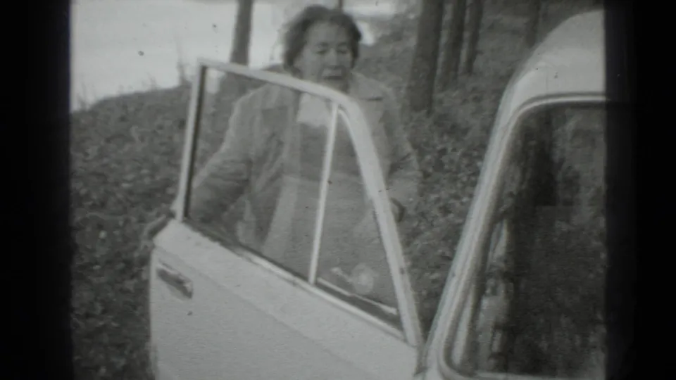 In a grainy black-and-white 8mm frame, a woman in a light coat stands by the open car door in Moscow, 1975.