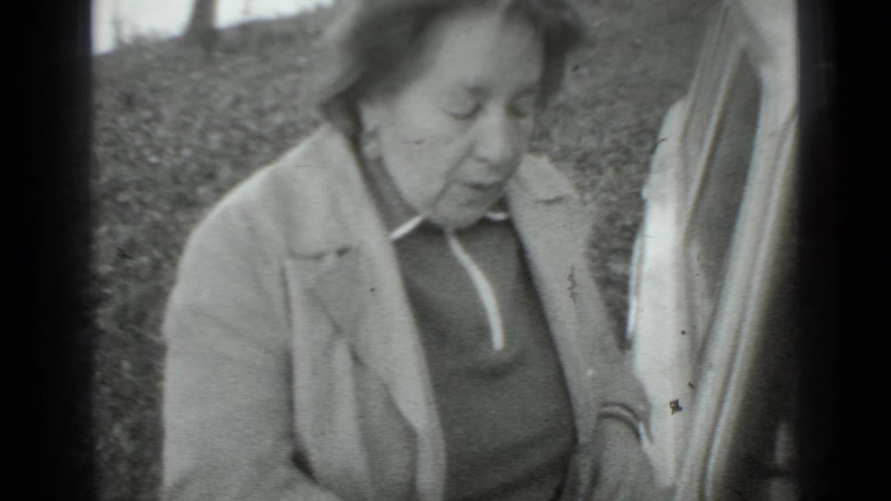Older woman in a light coat stands beside a car outdoors.