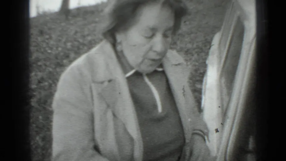 Older woman in a light coat and dark sweater stands beside a car in 1975 Moscow home-movie footage.