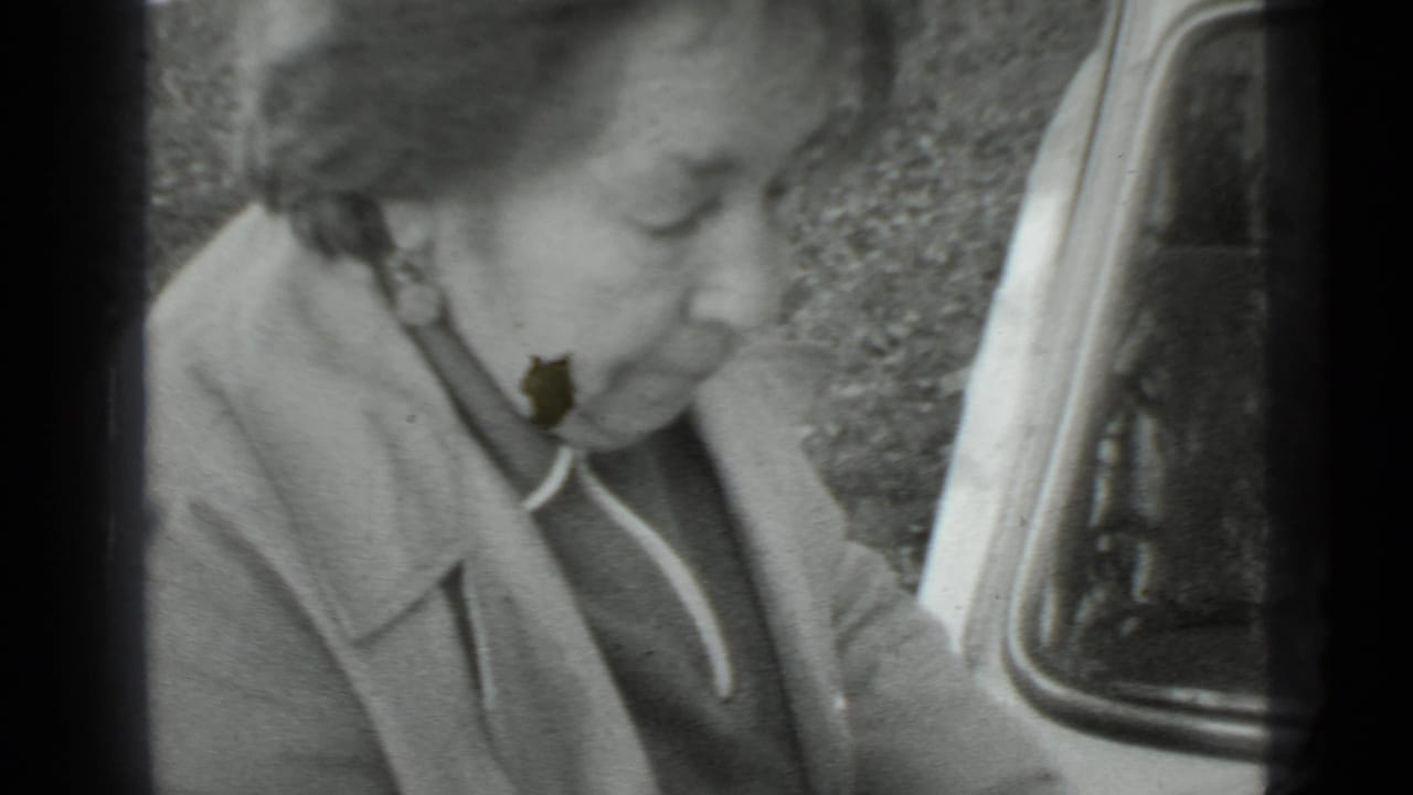 Woman in a coat bends beside a vehicle edge while working outdoors.