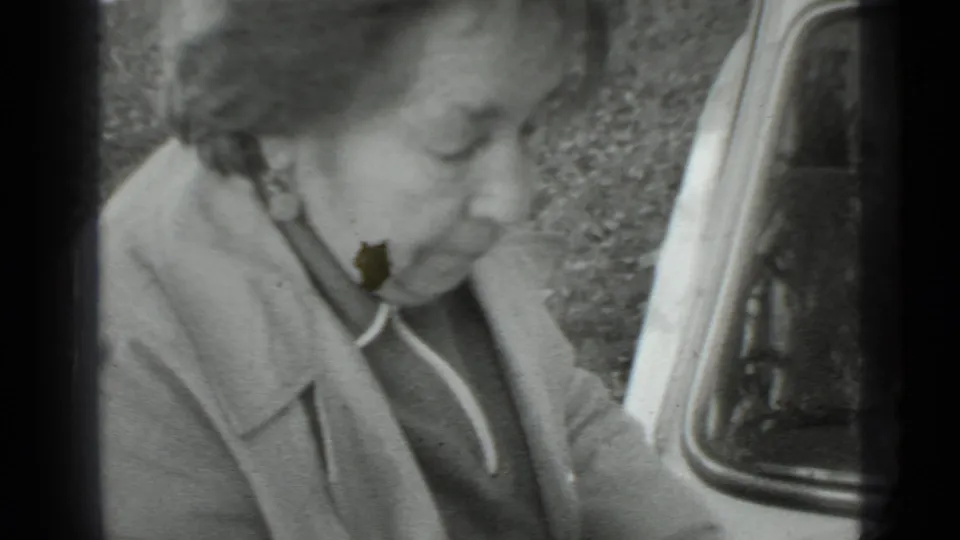 In Moscow 1975 home-movie footage, a woman in a grey coat bends near a vehicle edge during outdoor garden work.