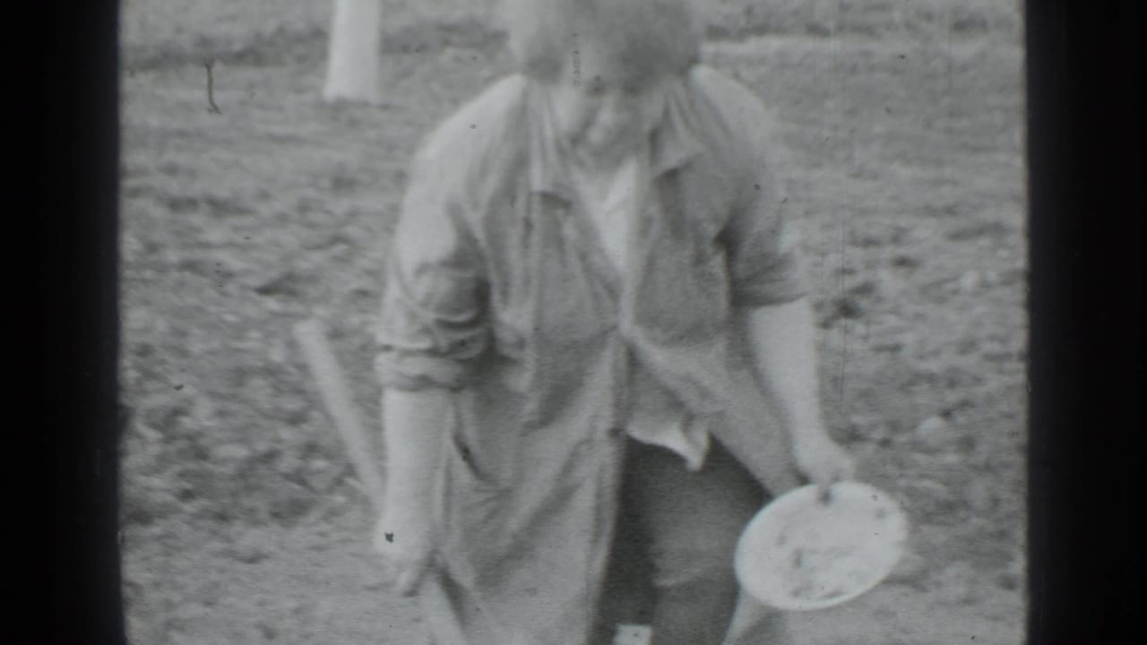 Woman stands in a garden holding a hoe and a round container.