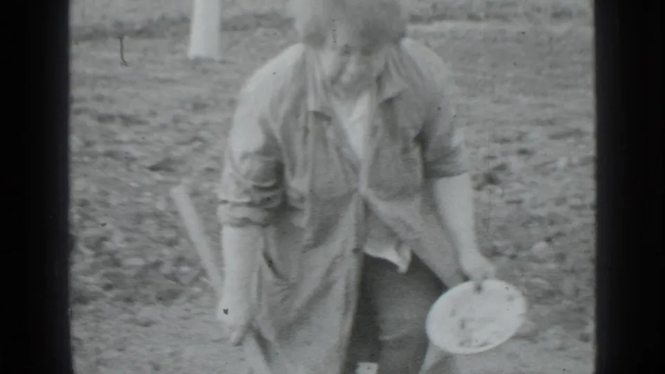 A woman in Moscow, Russia, 1975 stands on rough garden soil holding a hoe in one hand and a round container in the other.