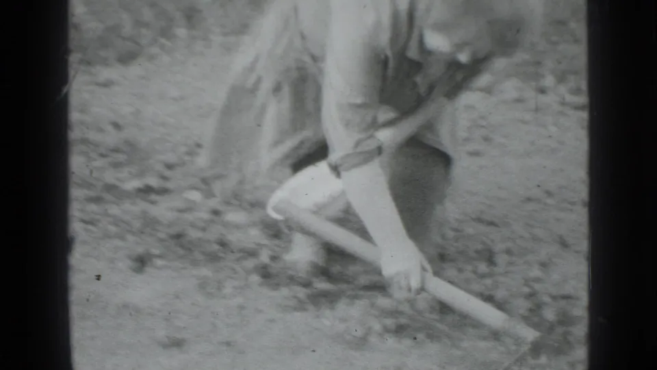 Close 8mm frame from Moscow 1975 showing a woman scraping bare garden soil with a long-handled hoe.