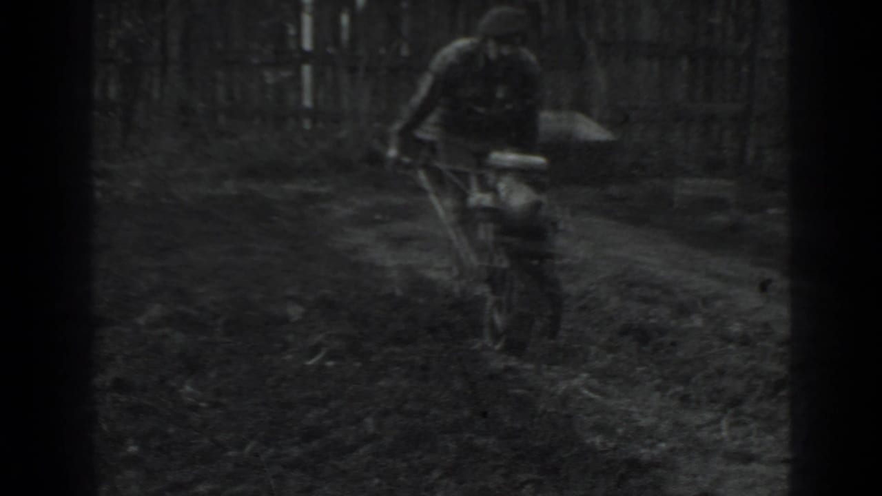 Man pushing a small tiller through a dark garden plot near a wooden fence.