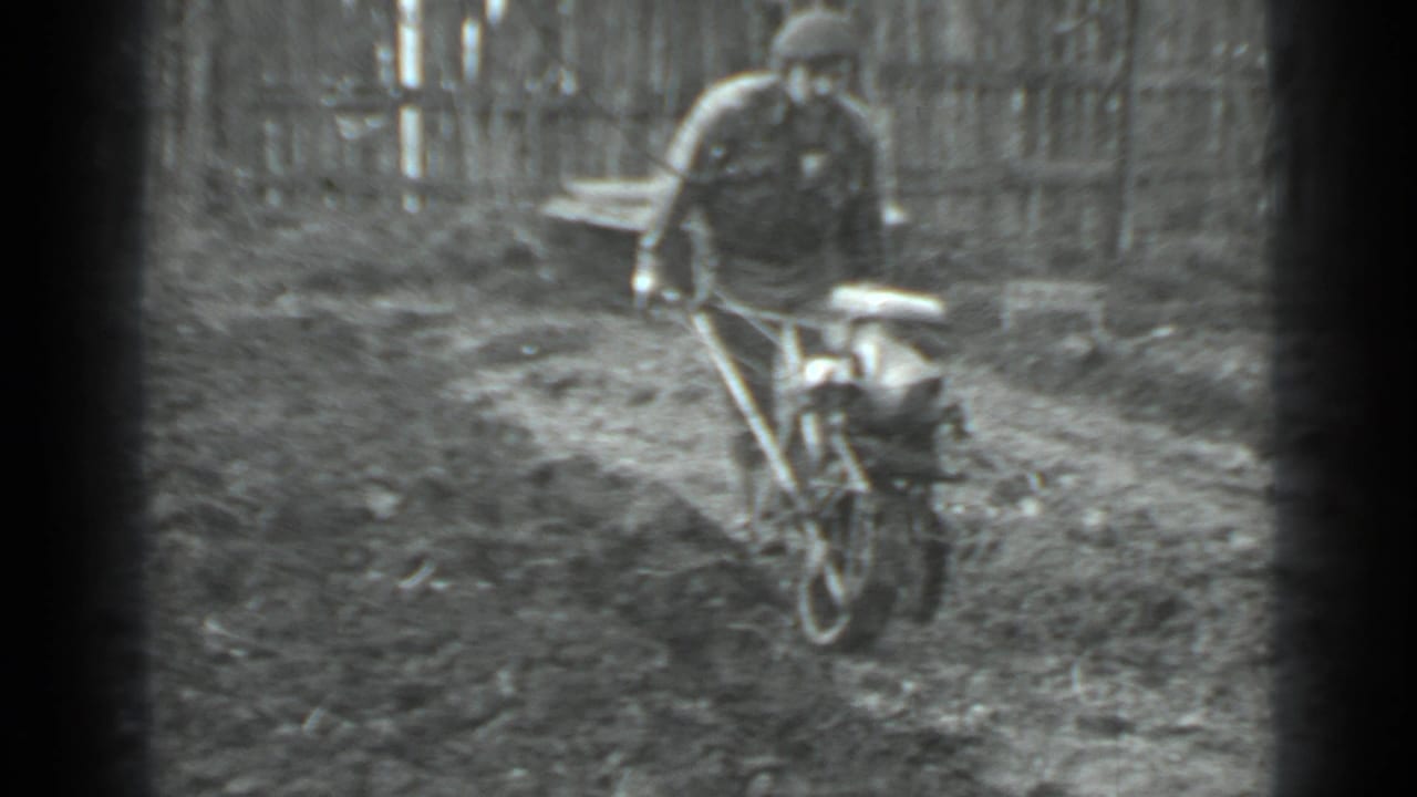 Man seen from behind operating a hand-pushed tiller in a fenced garden.