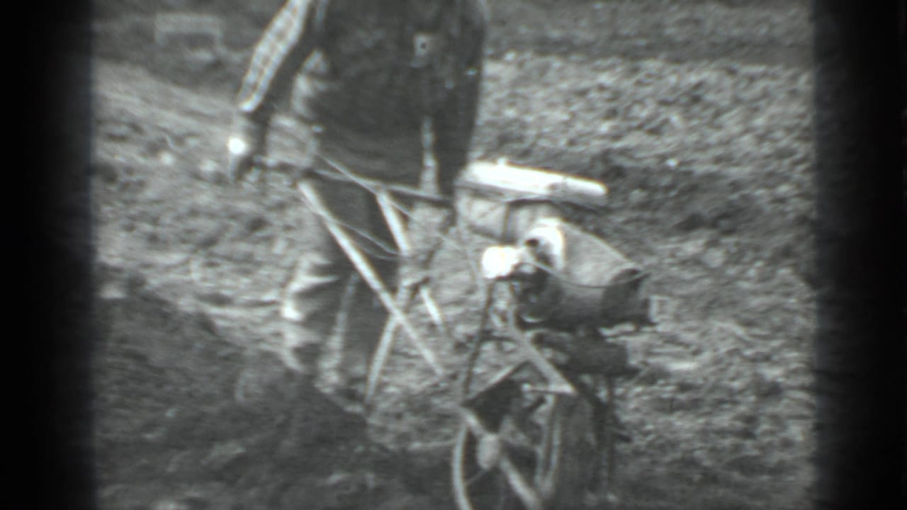 Archival 8mm frame from Moscow, Russia in 1975 showing a man working a small plow through uneven field soil.