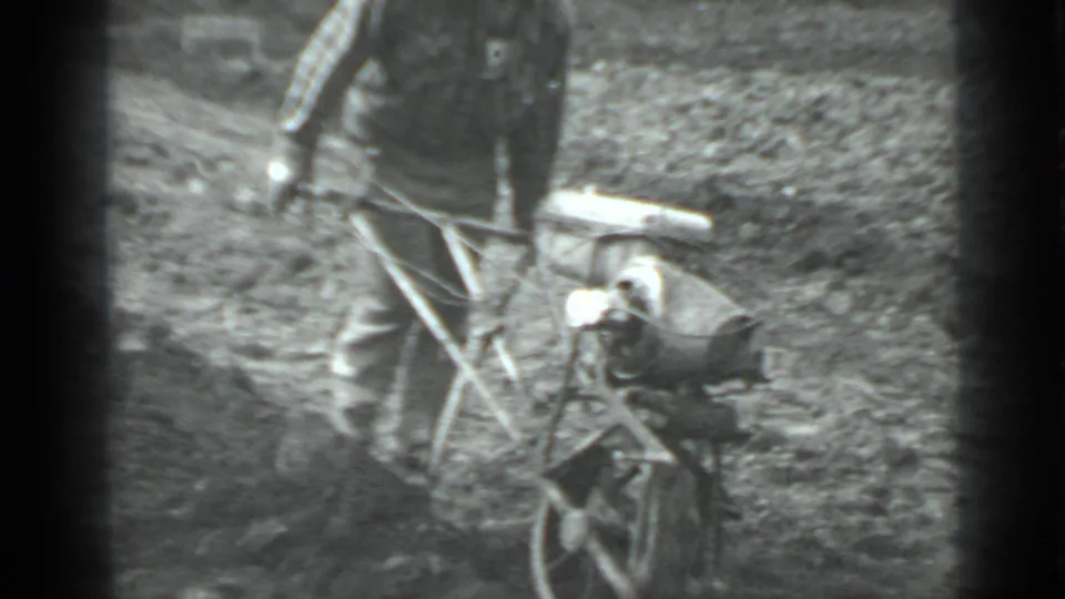 Archival 8mm frame from Moscow, Russia in 1975 showing a man working a small plow through uneven field soil.