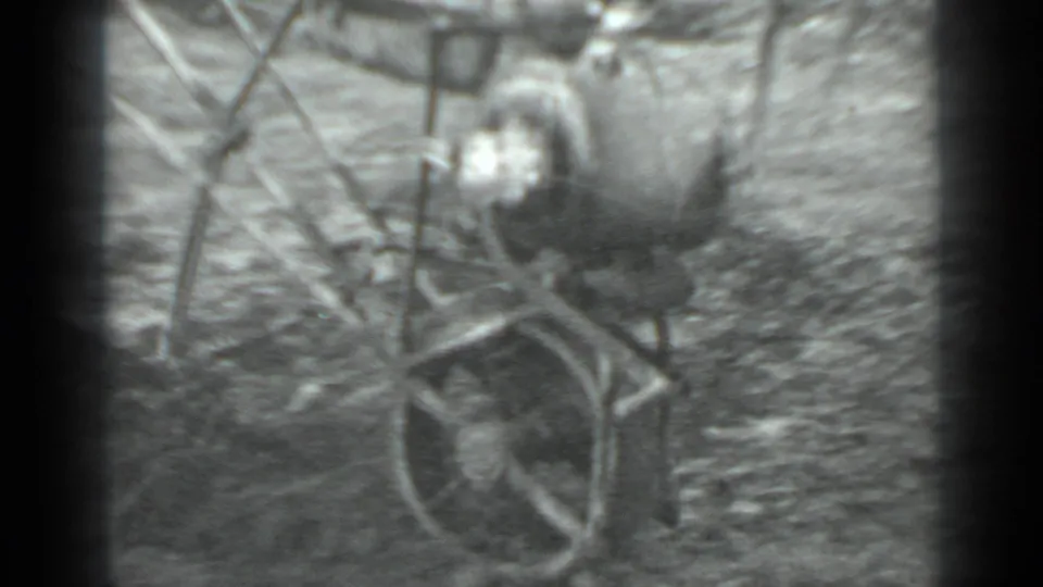 Close 1975 Moscow home-movie frame of a small walk-behind plow and its operator, with wheel and handles prominent against loose soil.