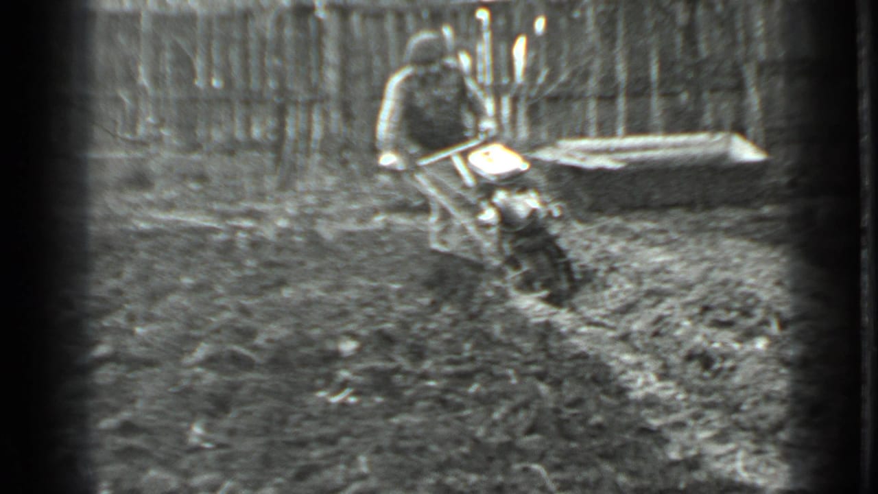The man continues wrestling the tiller through wet soil in the fenced garden, with soft black-and-white 8mm texture.