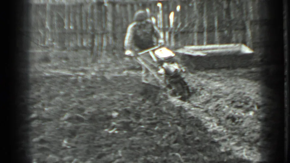 The man continues wrestling the tiller through wet soil in the fenced garden, with soft black-and-white 8mm texture.