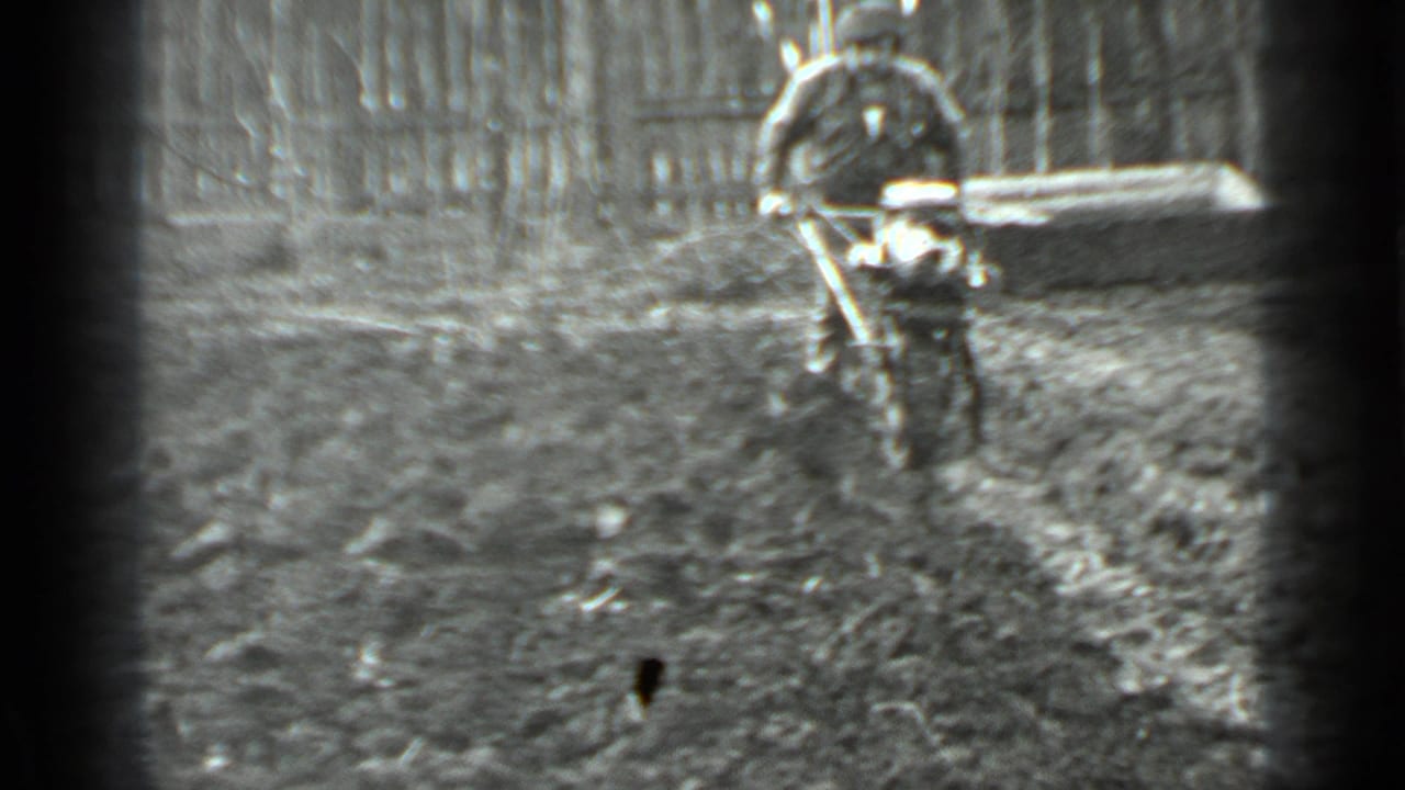 In 1975 Moscow 8mm footage, a man in dark clothing guides a small motorized cutter over uneven muddy ground beside a fence.