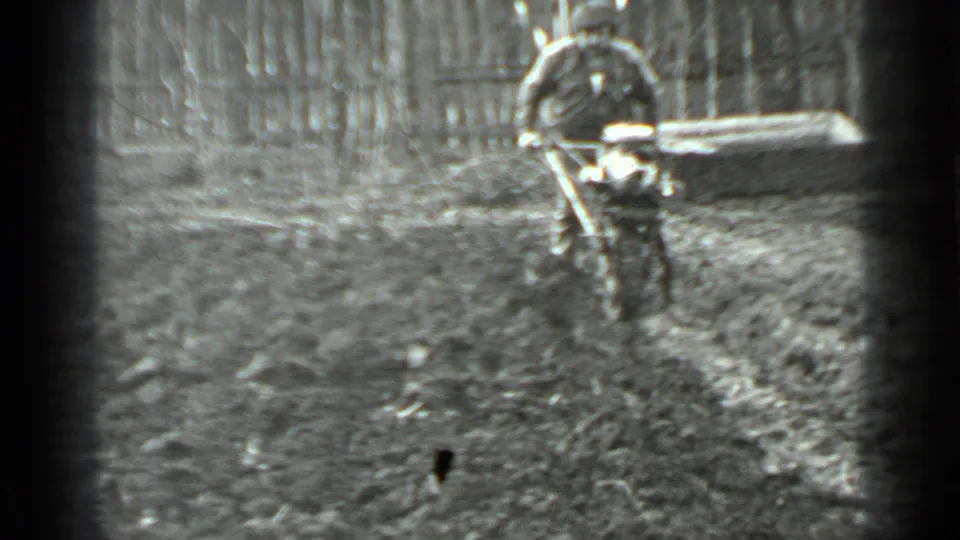 In 1975 Moscow 8mm footage, a man in dark clothing guides a small motorized cutter over uneven muddy ground beside a fence.