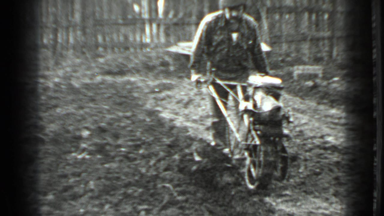 In 1975 Moscow home-movie footage, a man in a dark jacket steers a one-wheel tiller over uneven garden soil near a fence.