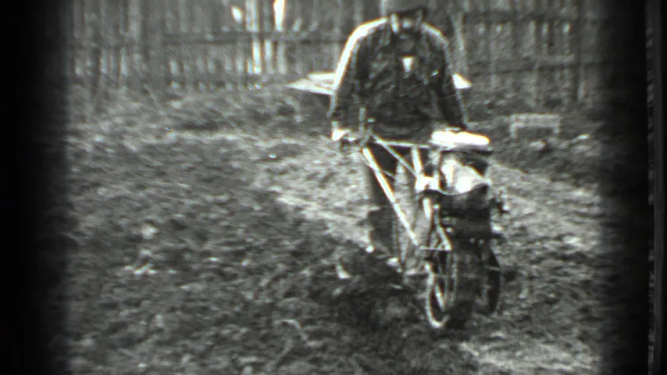 In 1975 Moscow home-movie footage, a man in a dark jacket steers a one-wheel tiller over uneven garden soil near a fence.