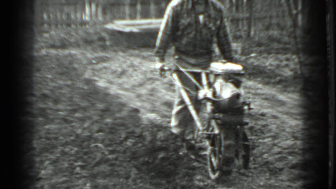 Man pushes a motorized tiller forward with a fence behind him.