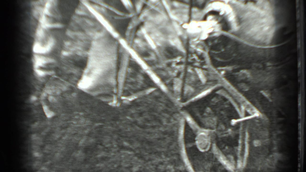 A rear view shows the man pushing the hand tiller across dirt, with branches and soft black-and-white 8mm grain around the frame.