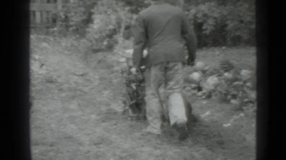 In 1975 Moscow home-movie footage, a man in a dark jacket and light trousers pushes a manual tiller through grass near plants and a fence.