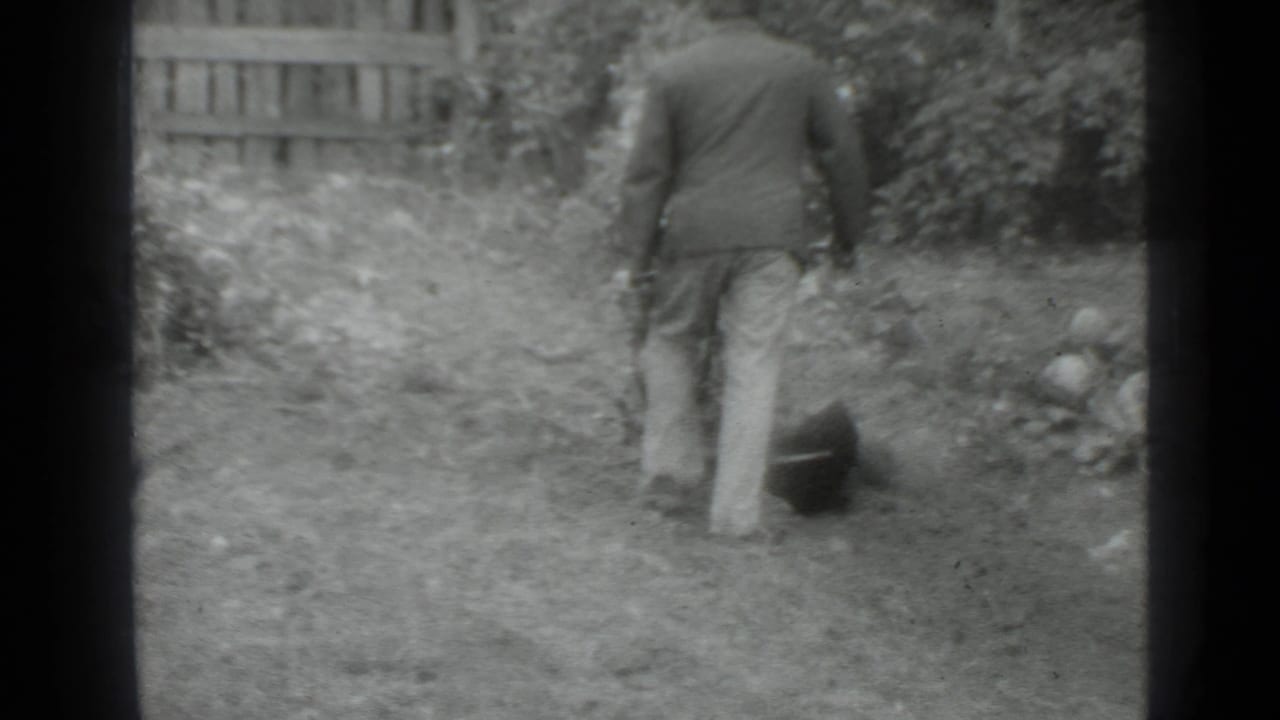 Man walks away across a grassy yard with a dark tool or bucket beside him.