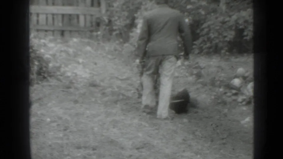 The man continues across the overgrown yard in the Moscow 1975 footage, with a dark garden object near his side and a wooden fence behind him.