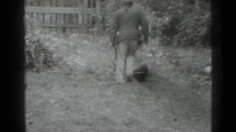 A rear view shows the man pausing or turning slightly in a fenced Moscow yard, with the manual garden tool visible near the ground.